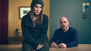 VICKY MCCLURE as Stella Tomlinson sitting on a desk in front of and JOHNNY HARRIS as Charles Stone.