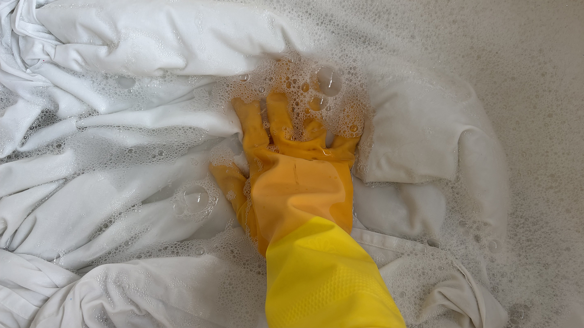 A hand wearing a yellow rubber glove washes a set of bed sheets in a bathtub filled with water and oxygen bleach