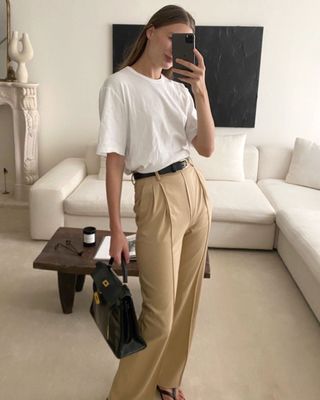 Die New Yorker Mode-Influencerin Christie Tyler posiert für ein Spiegel-Selfie in ihrem schicken Wohnzimmer und trägt ein weißes T-Shirt, einen schwarzen Gürtel, eine Vintage-Hermès-Tasche, hoch taillierte braune Bundfaltenhosen und flache schwarze Flip-Flops.