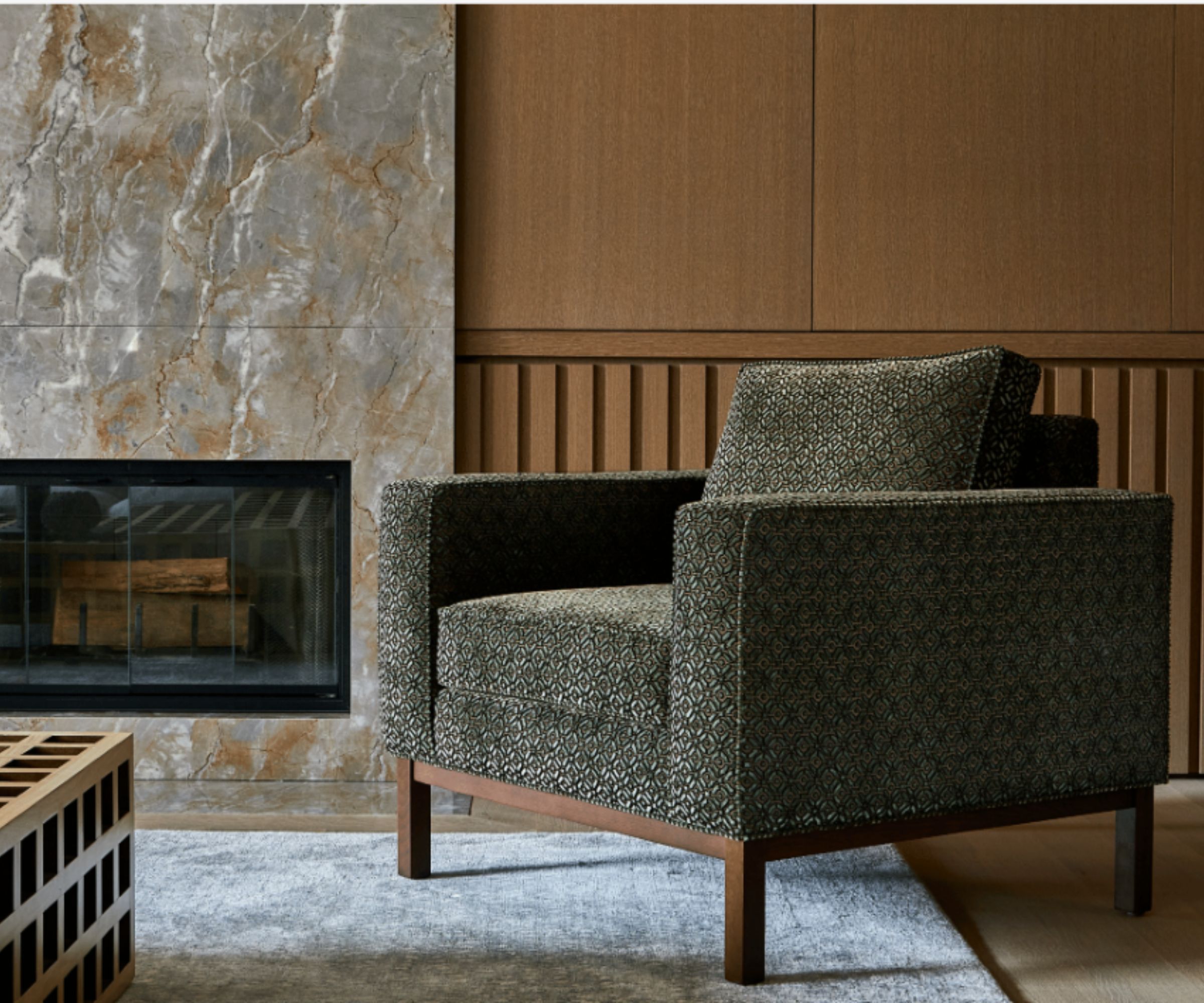 Close up of green upholstered armchair with wooden frame, neutral rug underneath, marble fireplace in background with wood panelled walls