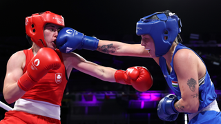 A female boxer (on the right) wearing blue kit is shown punching the face of another boxer wearing red kit (on the left.)