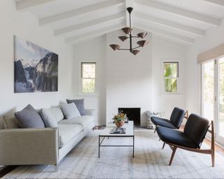 White living room with large rug, two armchairs facing gray sofa, white and black metal coffee table, chandelier, fireplace, artwork on wall, vaulted ceiling