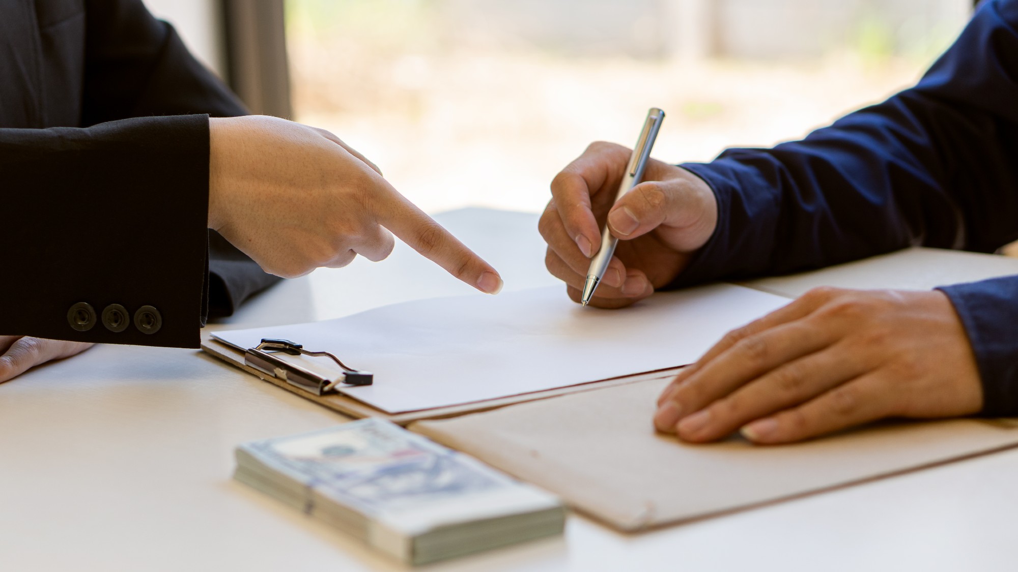 One person is signing a contract while another person is pointing to the correct place to sign on the document. A pile of money sits on the table next to them.