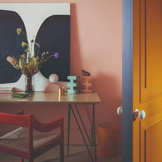 Pink-painted home office with a red chair at the desk and an art work with a vase of flowers on top of the desk