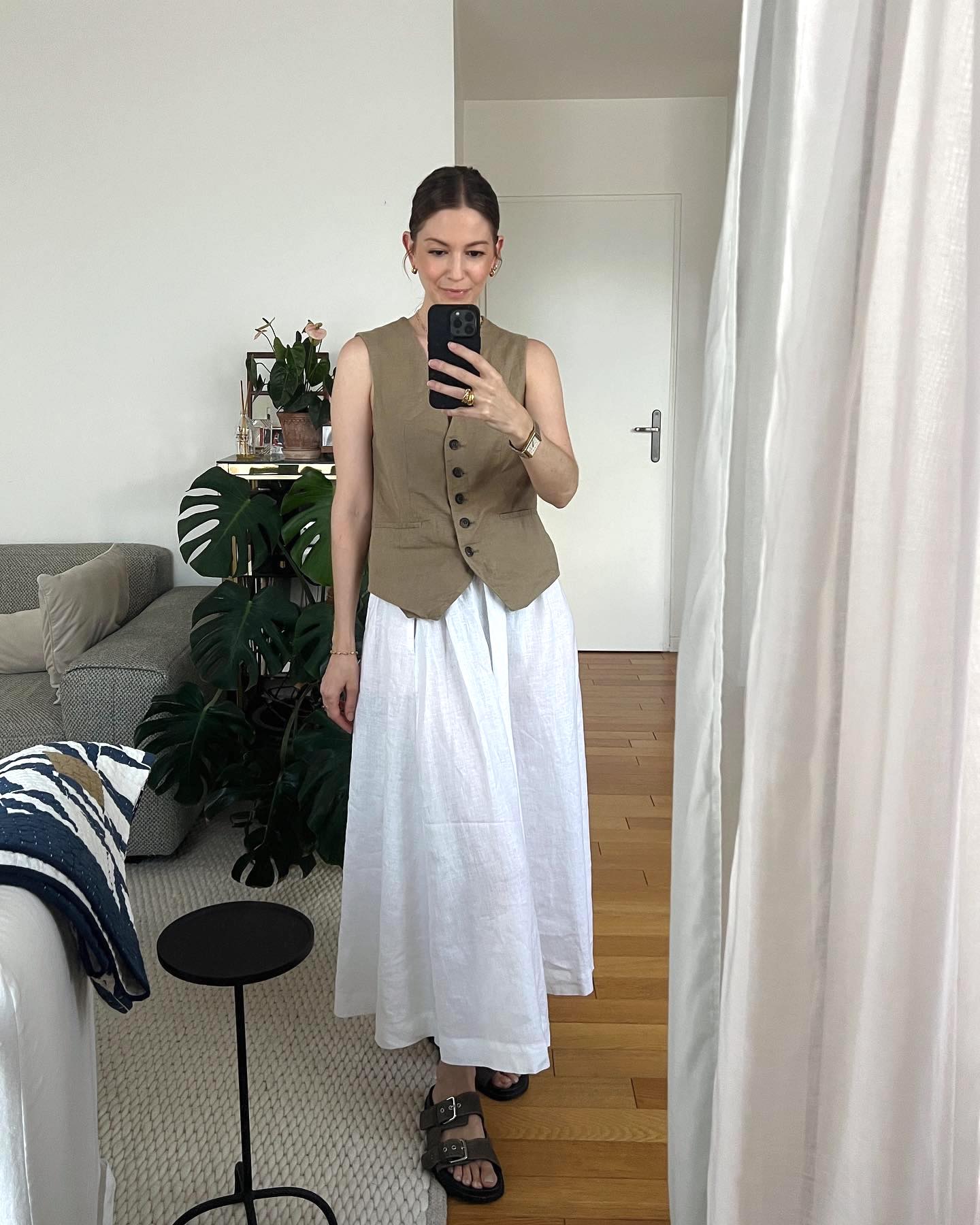stylish fashion influencer poses for a mirror selfie in her living room wearing a tan button-down vest top, full white skirt, and buckled slide sandals