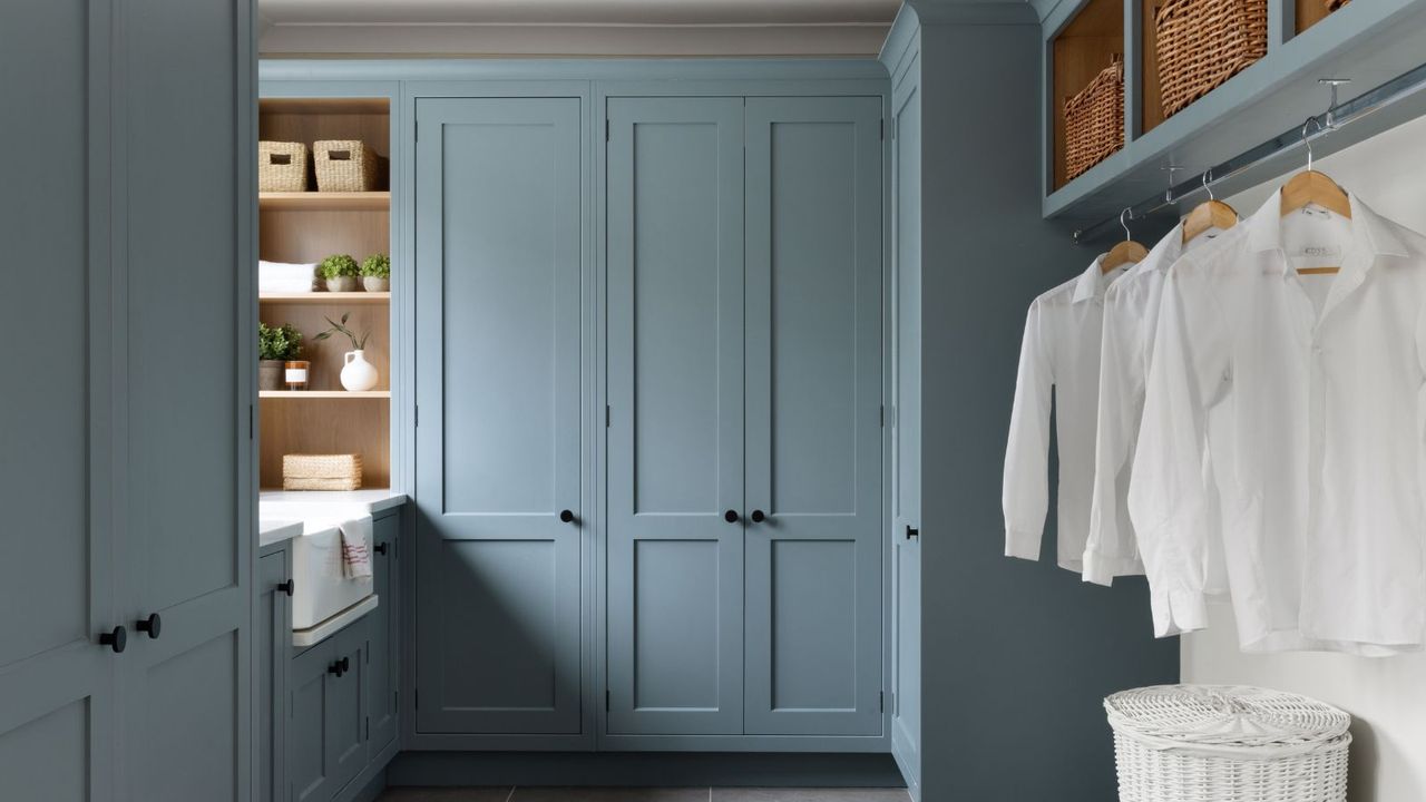 A large laundry room with tall blue painted built-in cabinets, a farm house sink under a window on the left hand side, and a clothes rail with pressed white shirts hanging from it on the right hand wall, baskets in storage holds above the rail