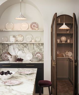 A white kitchen with an rounded alcove shelving unit, plates stacked on the shelves. To the right, an arched doorway into a pantry. Two milk glass fluted pendant lights hanging fom the ceiling.