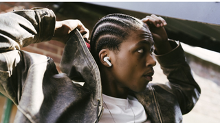 A man with cornrows wearing a white t-shirt and grey leather jacket models the Cambridge Audio Melomania A100 earbuds in white
