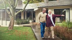 An older couple walk down the front walk outside their home, looking happy together.