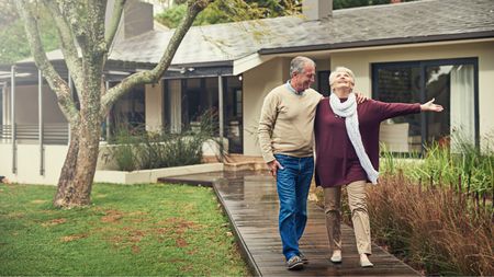 An older couple walk down the front walk outside their home, looking happy together.