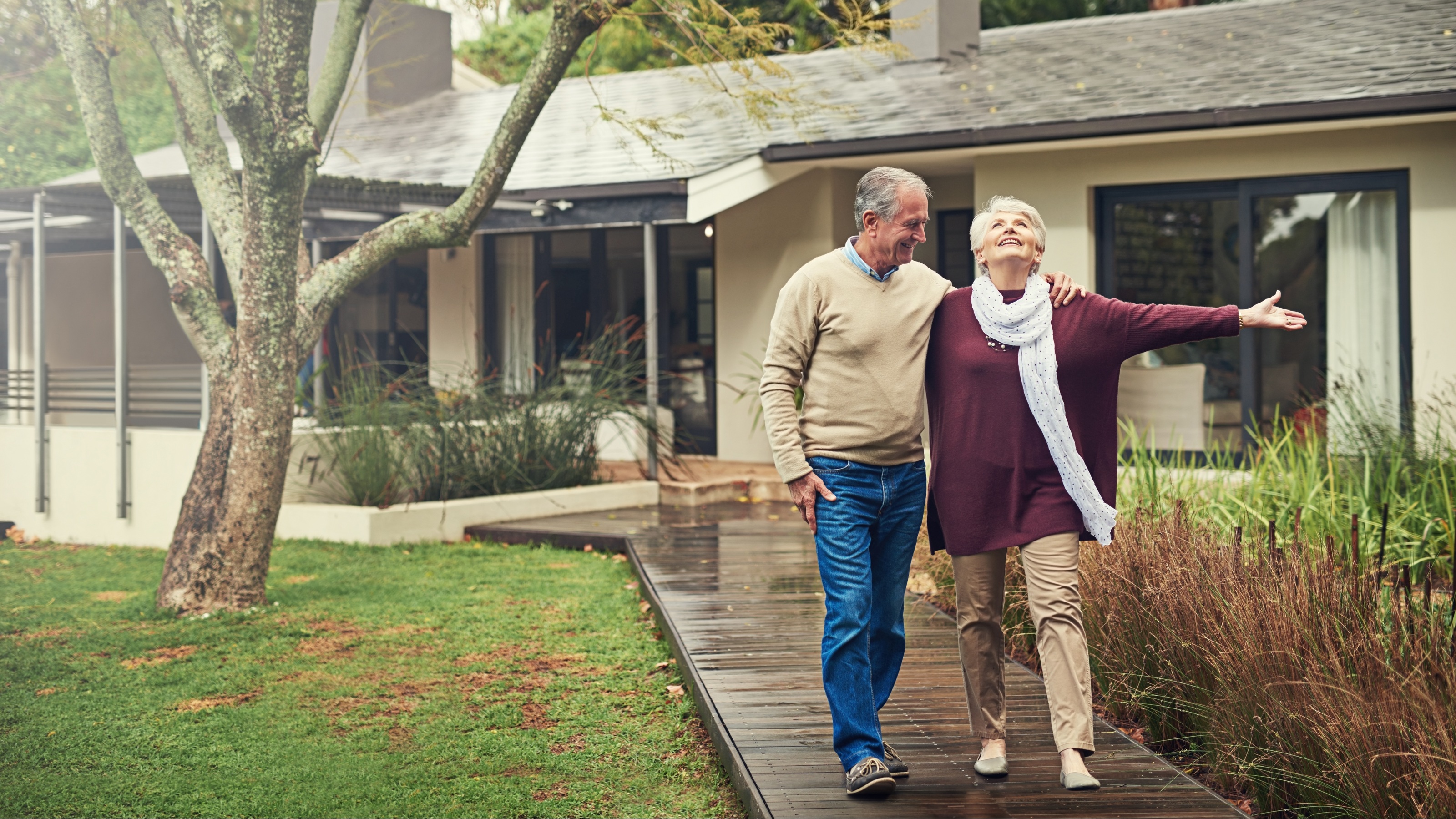An older couple walk down the front walk outside their home, looking happy together.