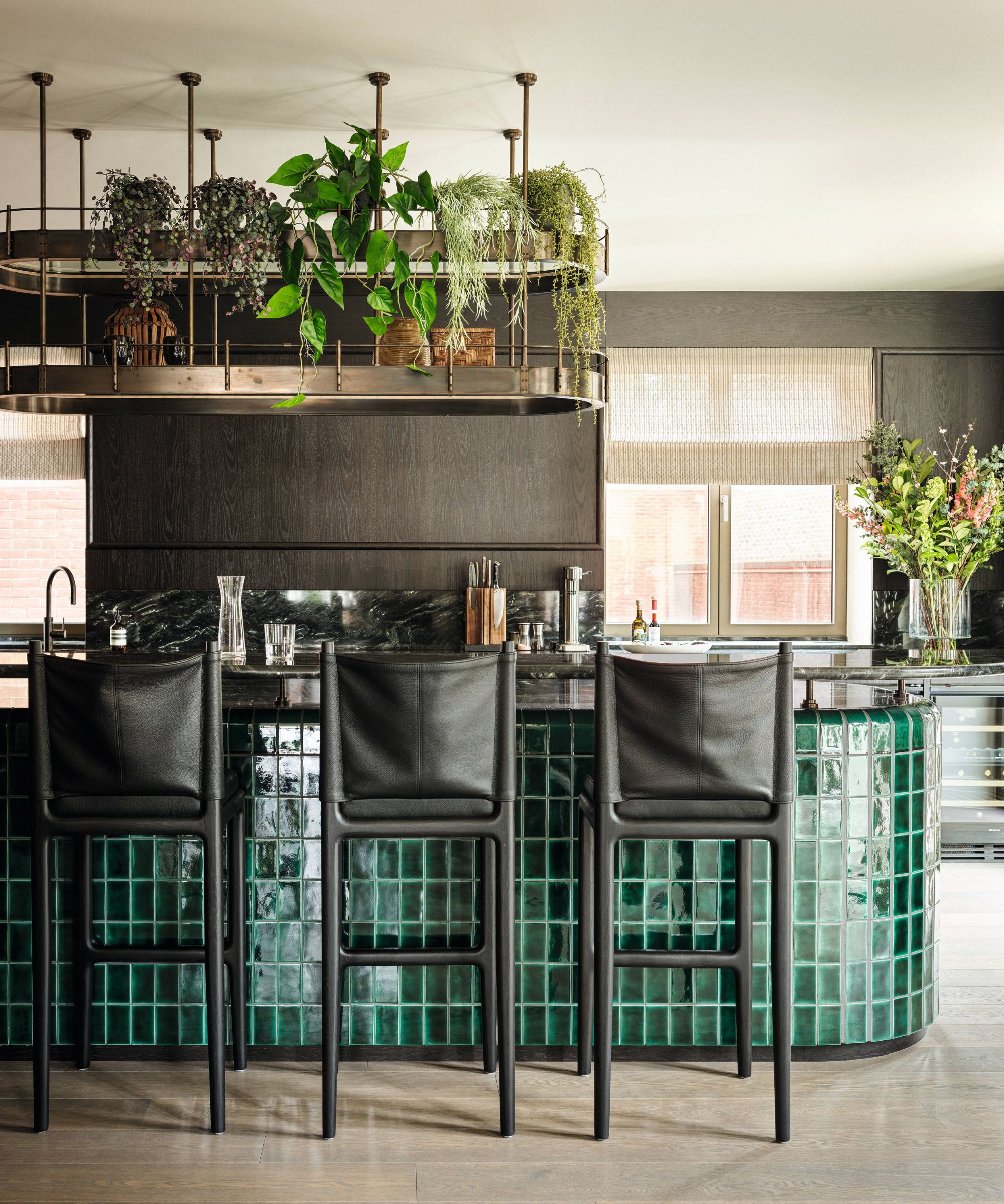 a contemporary gray and green kitchen with a hanging pot and plant rack, green tiled island with black wood and leather bar stools