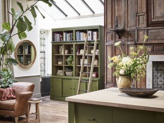 open plan kitchen with green kitchen island and dresser with ladder, rustic wood panelling, leather armchair, wooden floor, light and airy