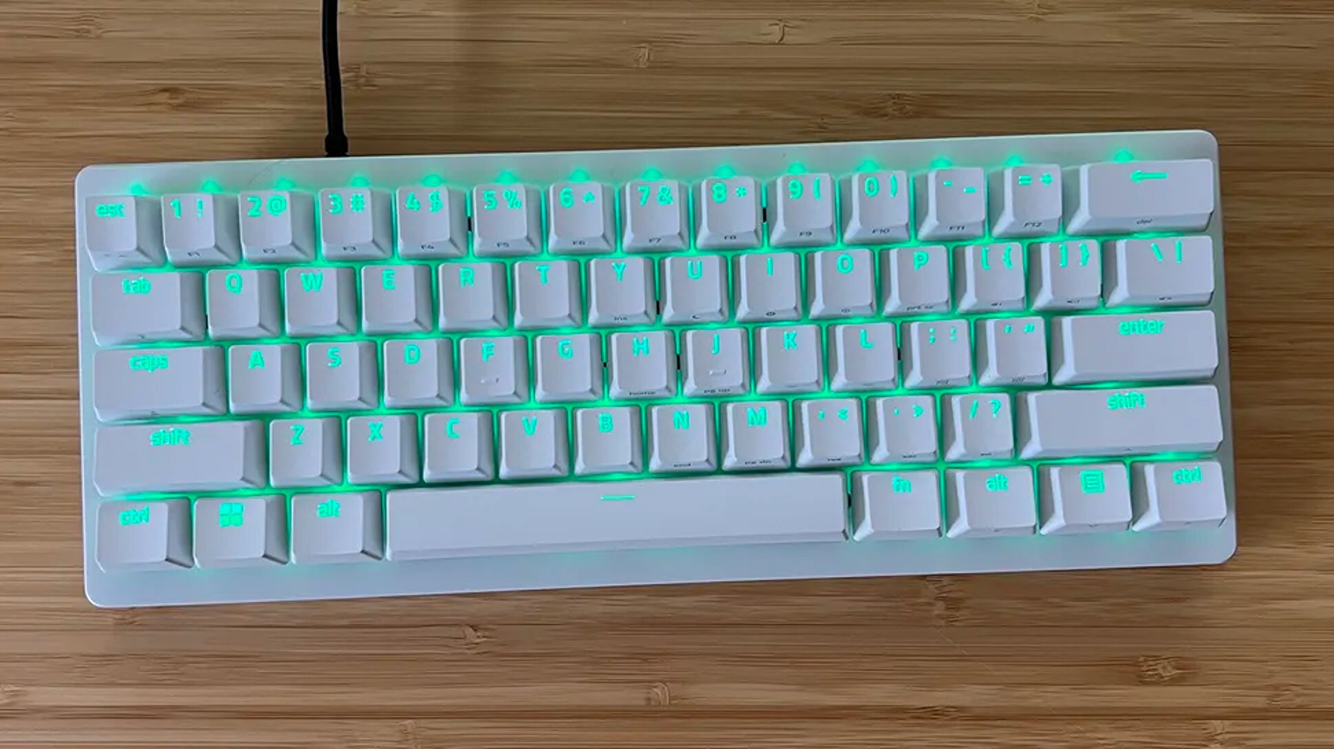 A compact white mechanical keyboard with glowing green backlighting sits on a light brown wooden desk, viewed from directly above in a top-down shot.