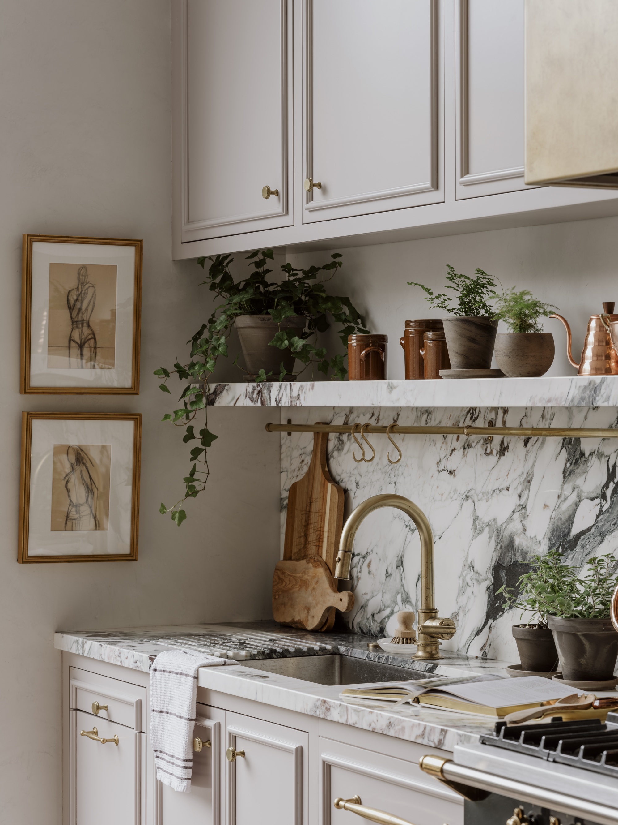 traditional style kitchen with marble countertop and backsplash, beige shaker-style cabinets, brass tapware, artwork hanging on wall, floating shelf styled with pots and small trinkets, and potted herbs
