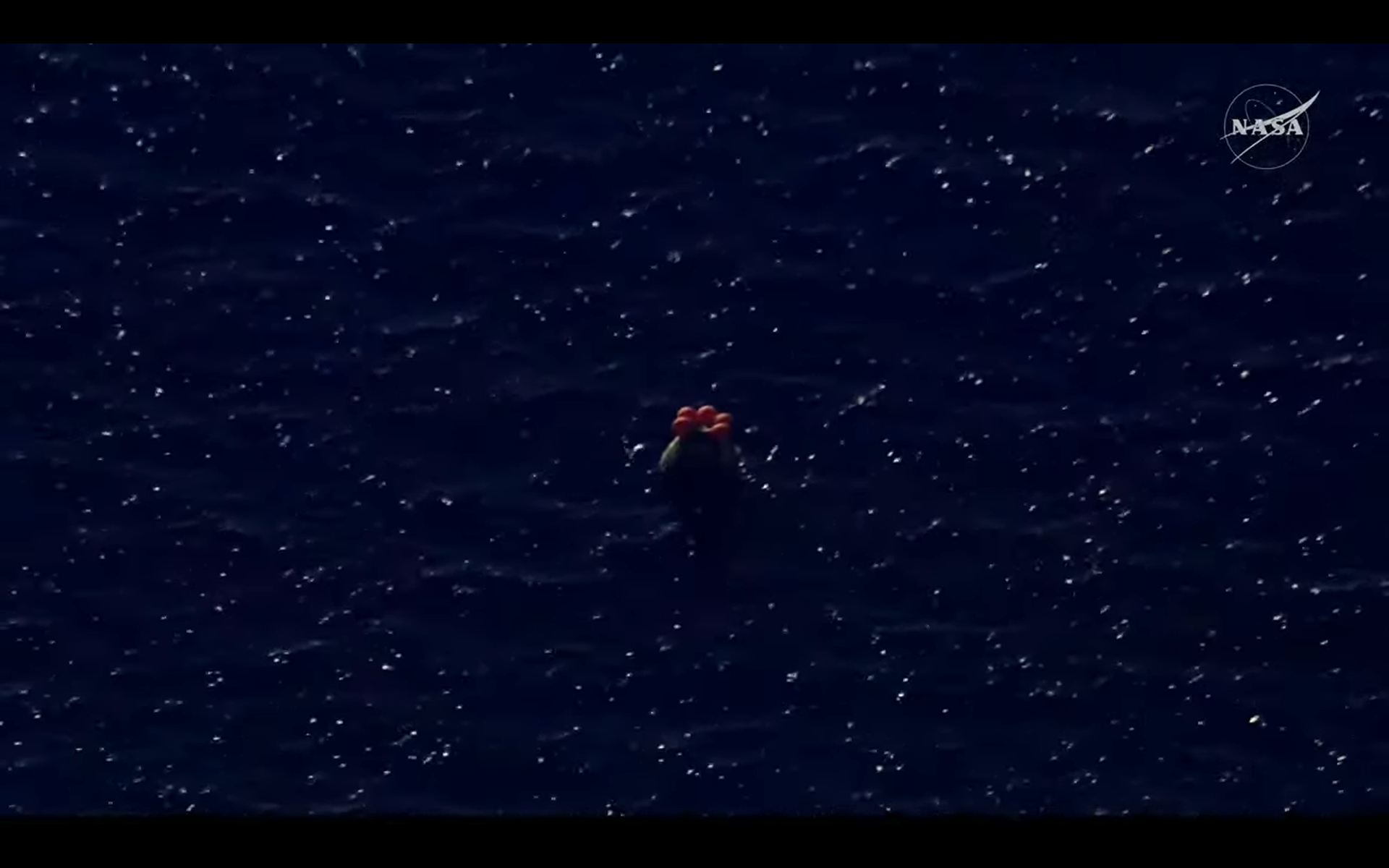 A round metal capsule with five orange balloons bobs on a dark blue ocean.