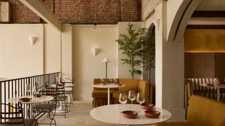A warmly furnished restaurant interior with wrought iron chairs topped by dark brown cushions, ochre banquettes, thriving plants, plaster butter walls, triangular sconces, and brick ceilings.