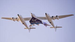 large airplane with a spaceship being carried underneath. the words "virgin galactic" are visible on the belly of the spaceship