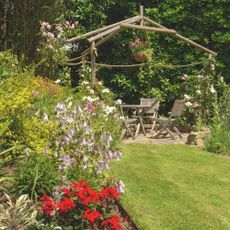 Lush garden borders next to an area laid to lawn, with a seating are with wooden chairs in the background