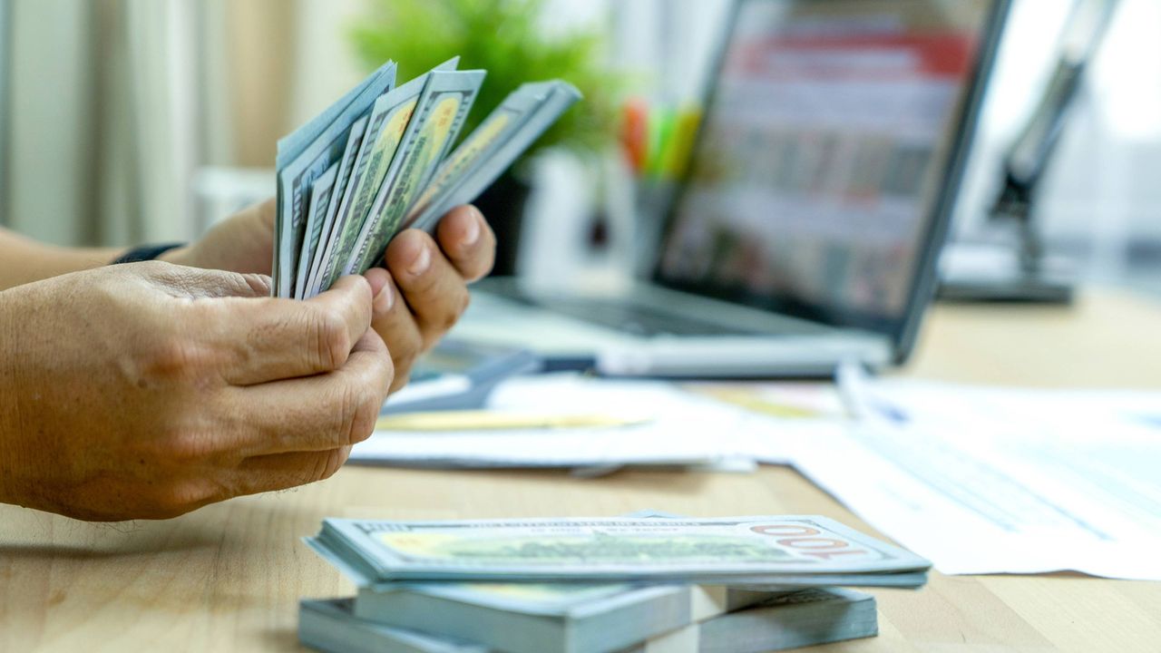 Hands hold a stack of money in front of a computer screen.