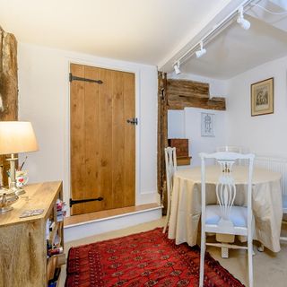 a small dining area with 4 seater circle dining table with cream tablecloth and white chairs, a red rug on the floor, wooden storage sideboard, wooden door and exposed timber on the walls
