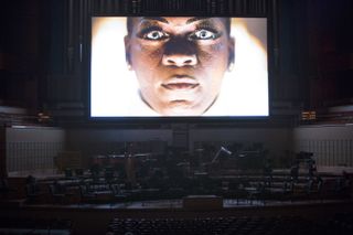 A screening of artist Yael Bartana's Inferno on a large screen. On the screen is the face of a black individual with blue eyes