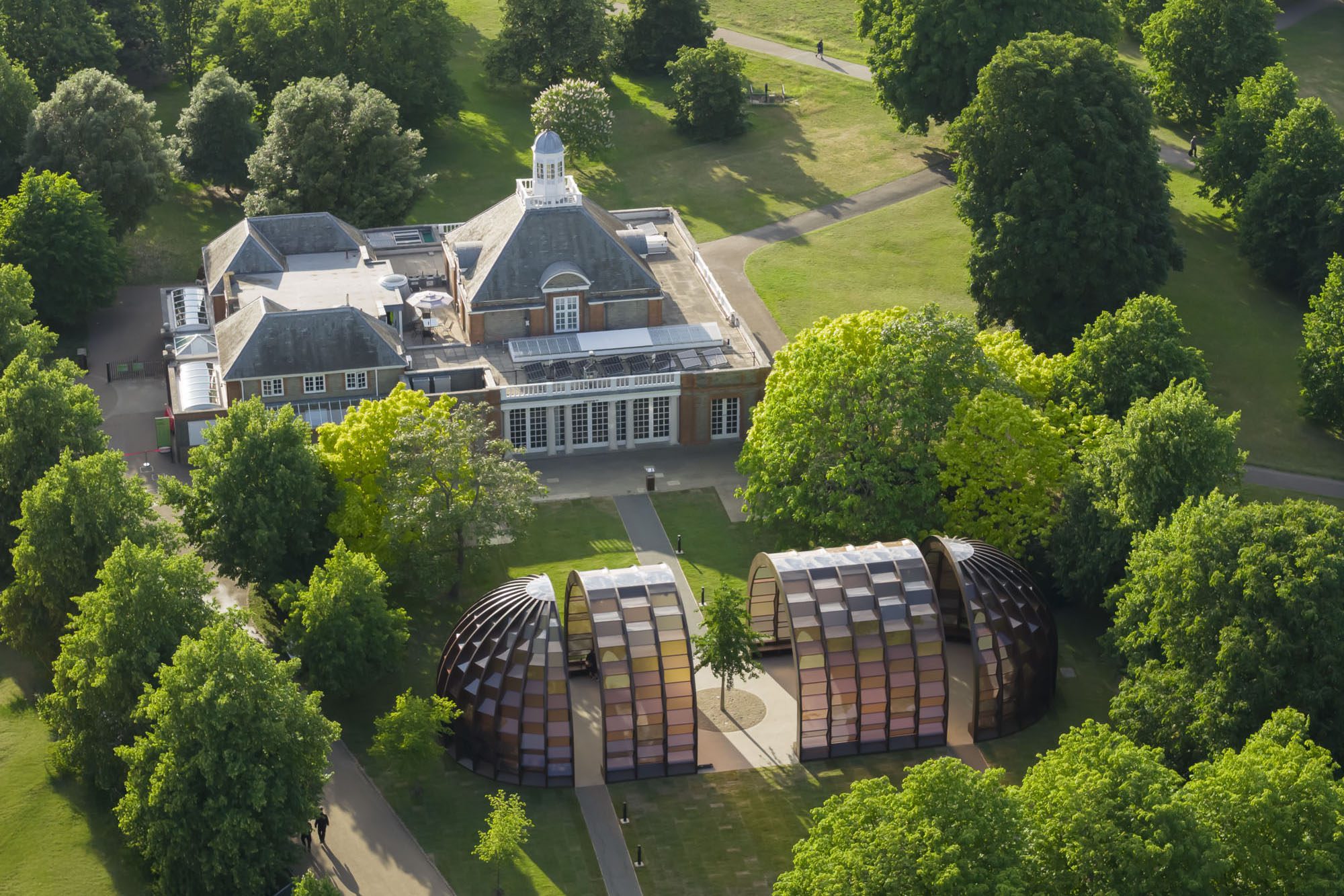 An aerial view of a contemporary architecture pavilion features lush vegetation and a beautifully preserved brick, glass, and stucco-style historical pavilion sitting next to it.