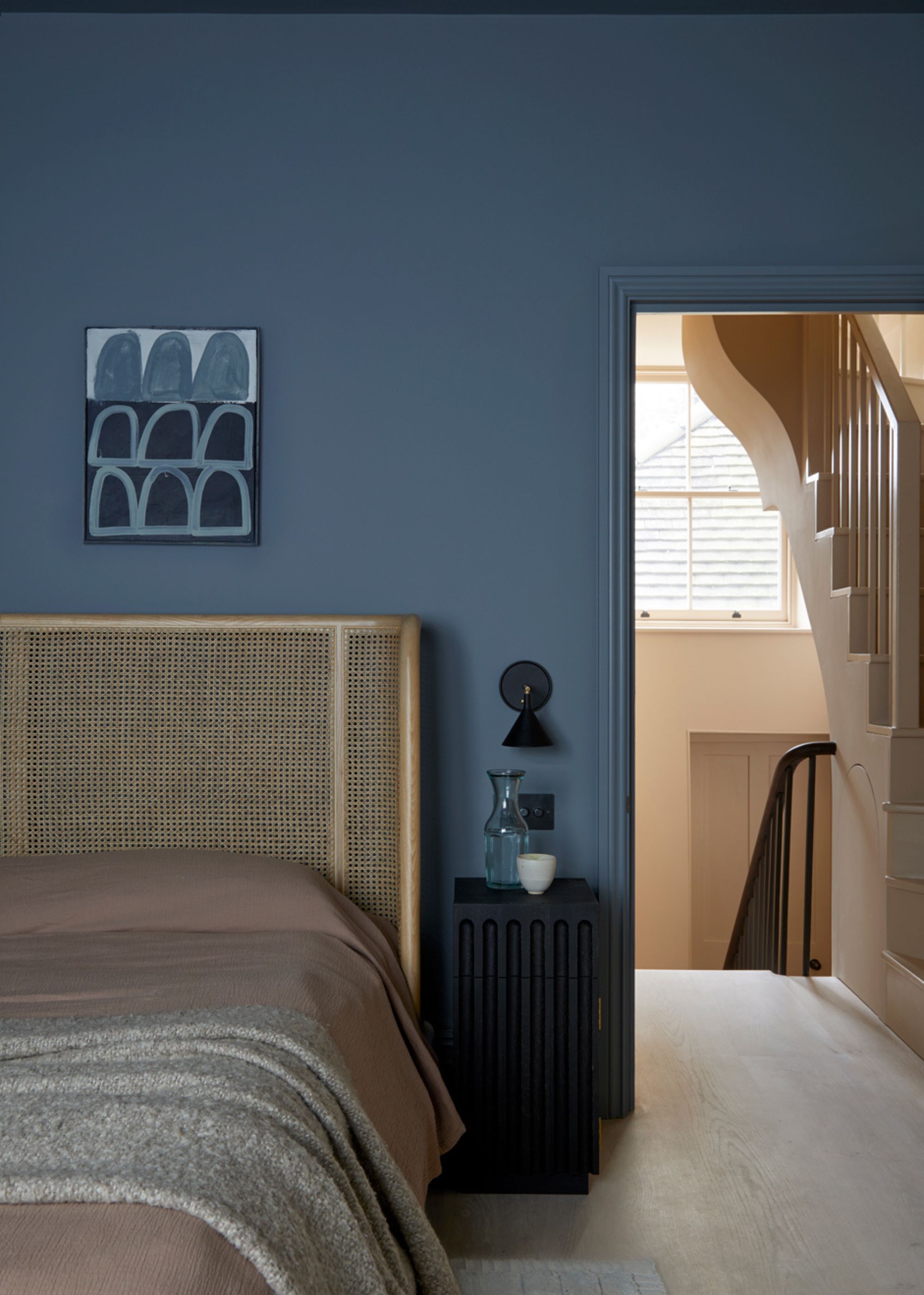 A bedroom with dark blue-gray walls, a rattan bed headboard with taupe bedding, and a gray throw. A doorway leading into a hallway painted plaster pink.
