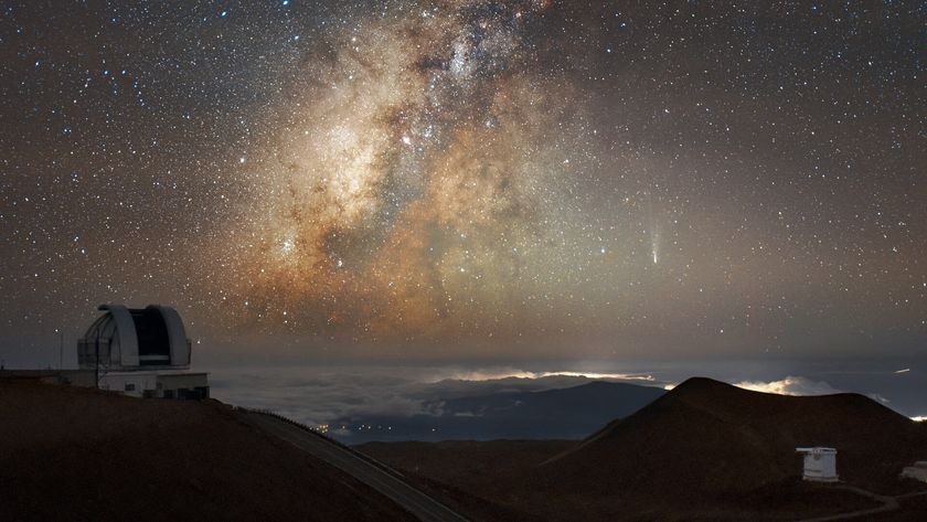 The Milky Way glows orange and yellow amidst the stars in the night sky, with a streak of white at the right side of the image where comet Lemmon moves across with a domed observatory and mountains silhouetted at the bottom