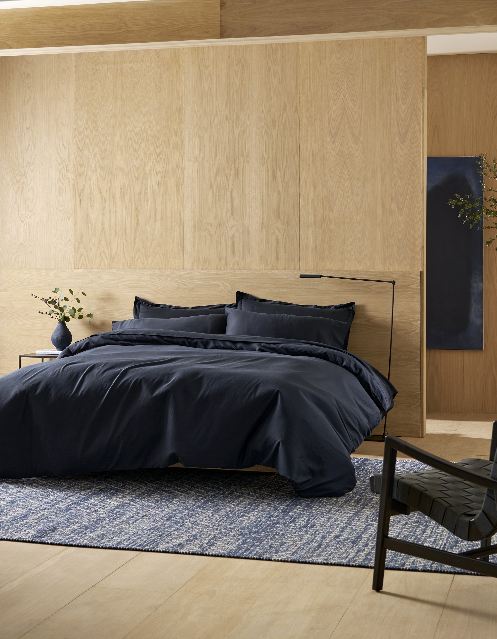 Cozy Earth soft wash cotton bedding in eclipse color in a room with timber walls and ceiling and floors, a blue rug, black armchair, and a false wall with a hallway behind with blue artwork
