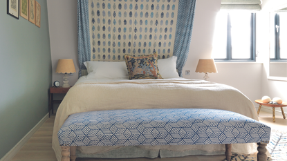 Double bed with blue curtain at the head of the bed and ottoman at the base, pale wooden floor and turquoise and white walls