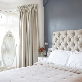 Blue and white bedroom with standing mirror, upholstered headboard and cream curtains