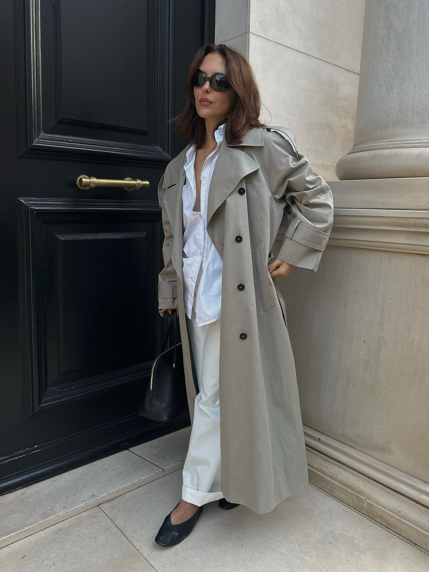 Style influencer Debora Rosa poses in a trench coat, white button-down shirt, whit cuffed pants, and black V-cut ballet flats.