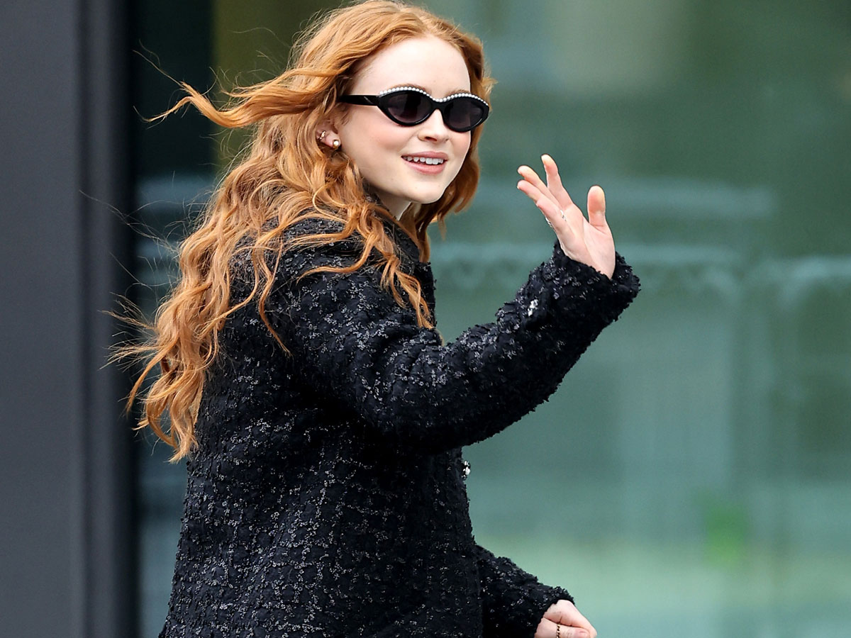 Sadie Sink wearing oval sunglasses and waving at the camera