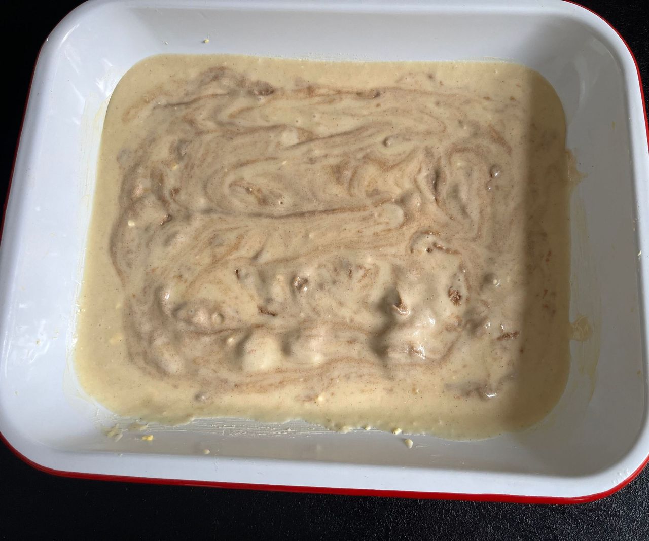 A white enamel dish with a red rim, with cinnamon swirled cake batter inside.