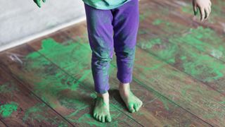 Child with green paid on hands and feed stood on hardwood floors with paint surrounding.