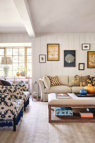 living room with white shiplap walls, beige couch and upholstered coffee table, rug, navy and beige print armchairs, artwork