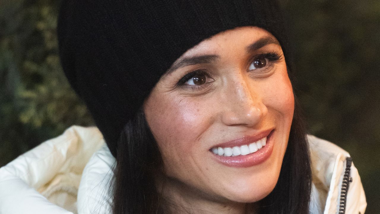 Meghan Markle smiling and wearing a black winter hat and white puffer coat