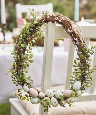 An egg shaped twine wreath with green leaves wrapped around it and pastel colored pink, blue, and yellow eggs layered at the bottom of it hanging on a white chair in front of a white table with decorations