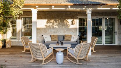 terrace patio off the back of a stone building with white pillars and decking patio area with wooden furniture, rocking chairs and a small blue side table