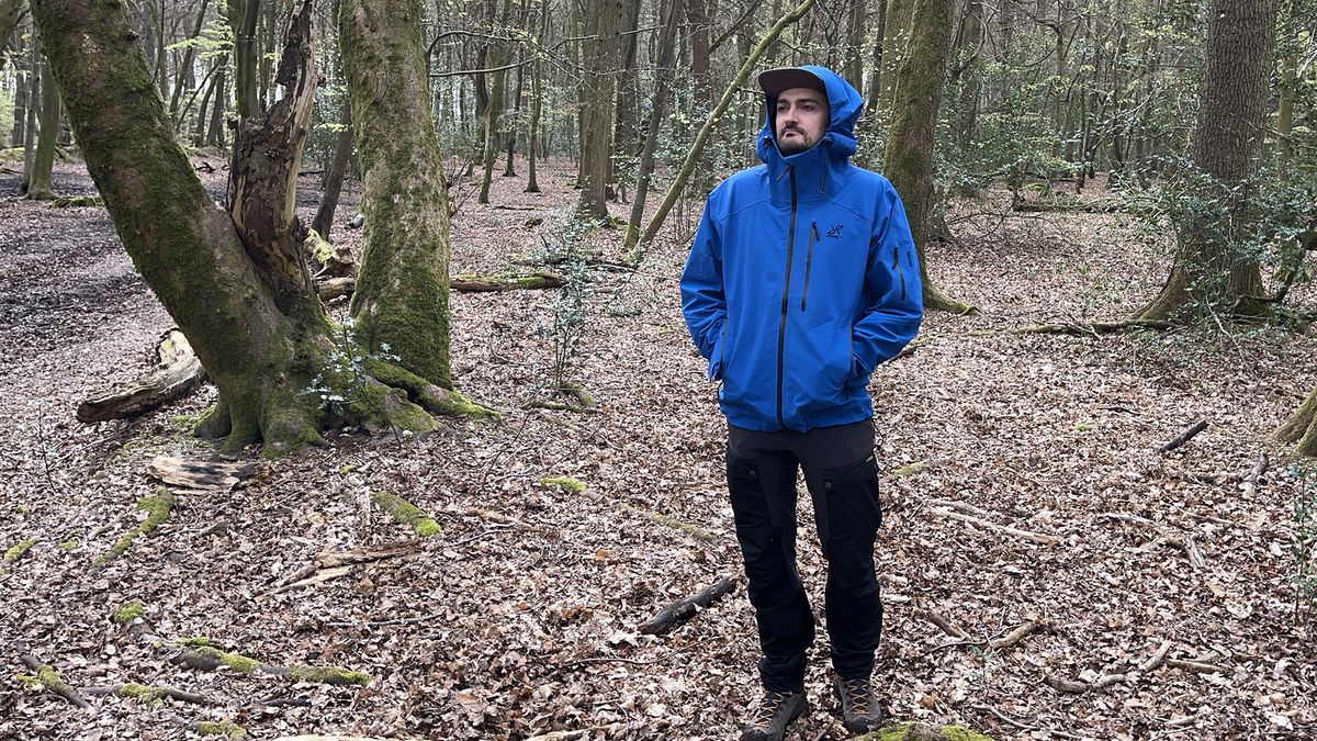 A man stands in a forest wearing a blue Revolution Race Aphex Pro waterproof jacket.