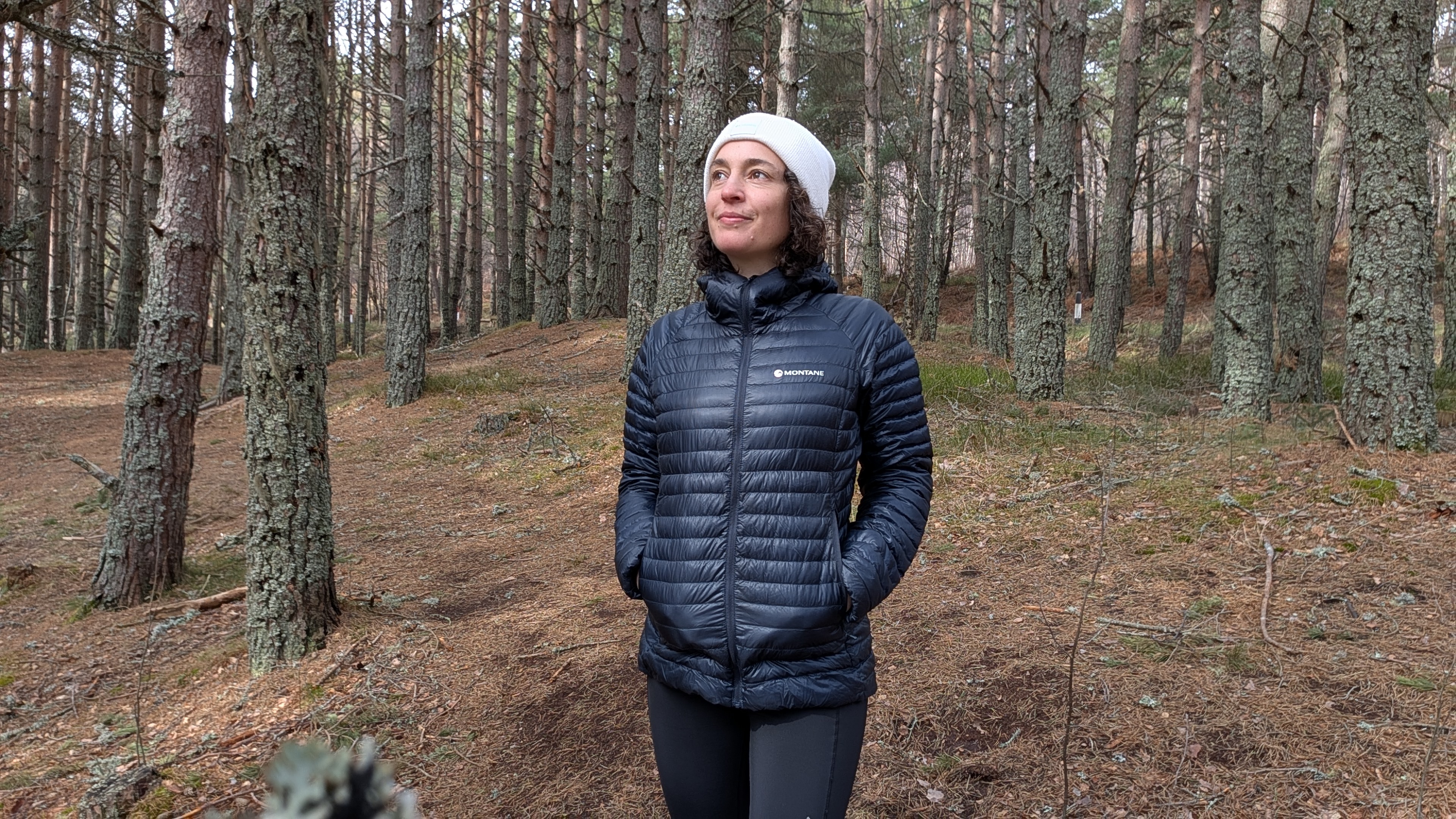 A woman in a forest wearing a dark blue down jacket and white hat with her hands in her pockets