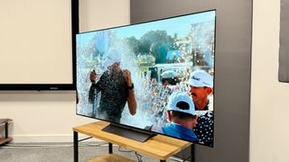 65-inch LG C4 TV photographed at an angle on a wooden stand. On the screen is an image of a golfer being sprayed with champagne.