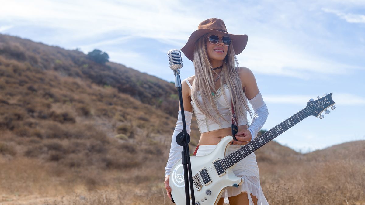 Orianthi stands at the mic in a still from the video for Some Kind of Feeling; she wears all white, shades and a wide-brimmed hat and is playing her white signature PRS electric.