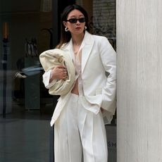 Woman wears white oversize blazer, sheer top, and white trousers white leaning against wall and holding off-white clutch handbag.