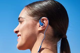 This image shows one of the best wired headphones for cycling, the JBL Endurance Run pair. The are in blue and on an ear of a woman who is side on to the camera facing left, with her dark hair in a low ponytail