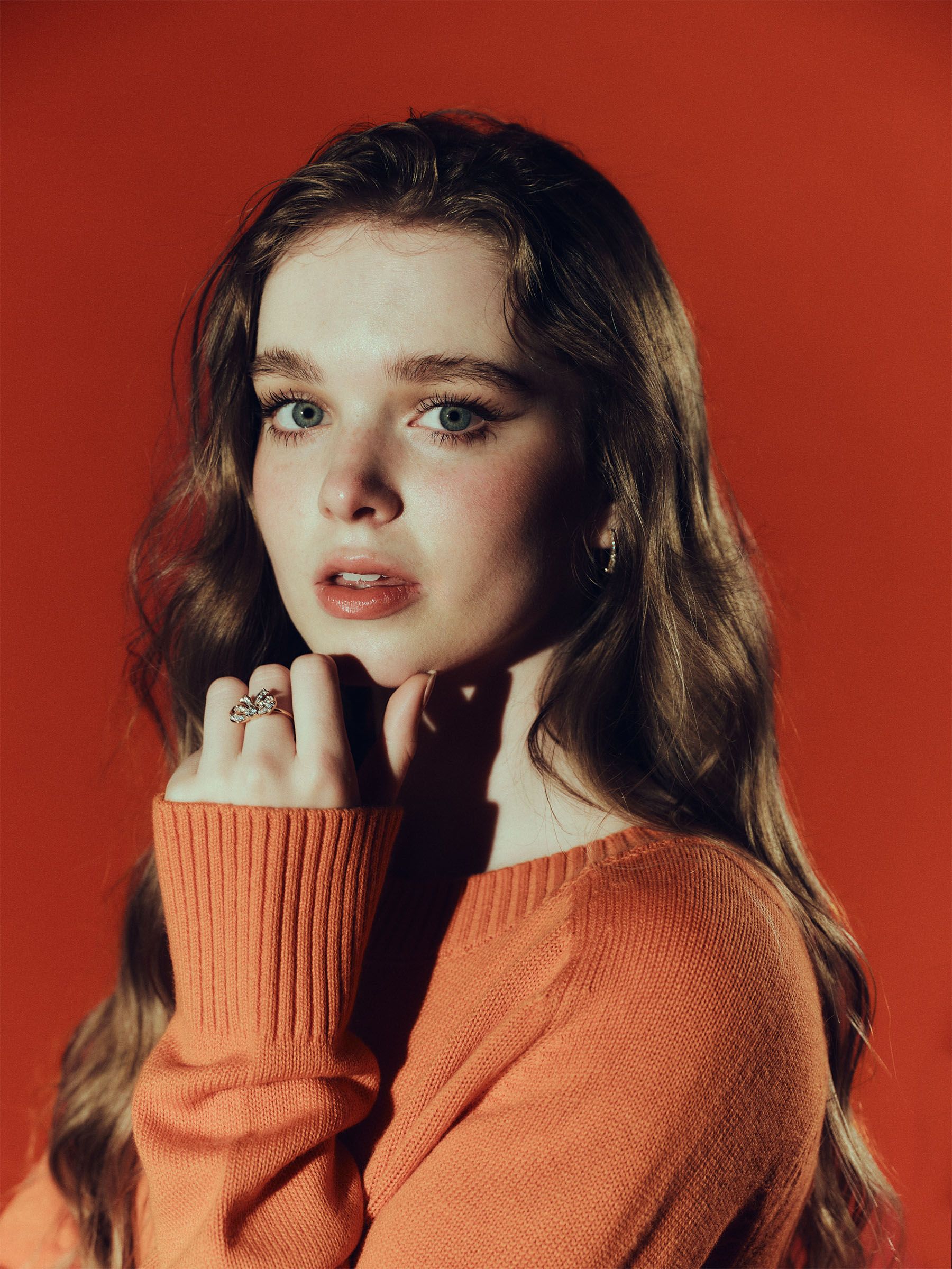 Carrie actress Summer Howell photographed by Jonny Marlow. She is looking to camera with her hand resting under her chin. She has on an orange sweater and a diamond bow ring.