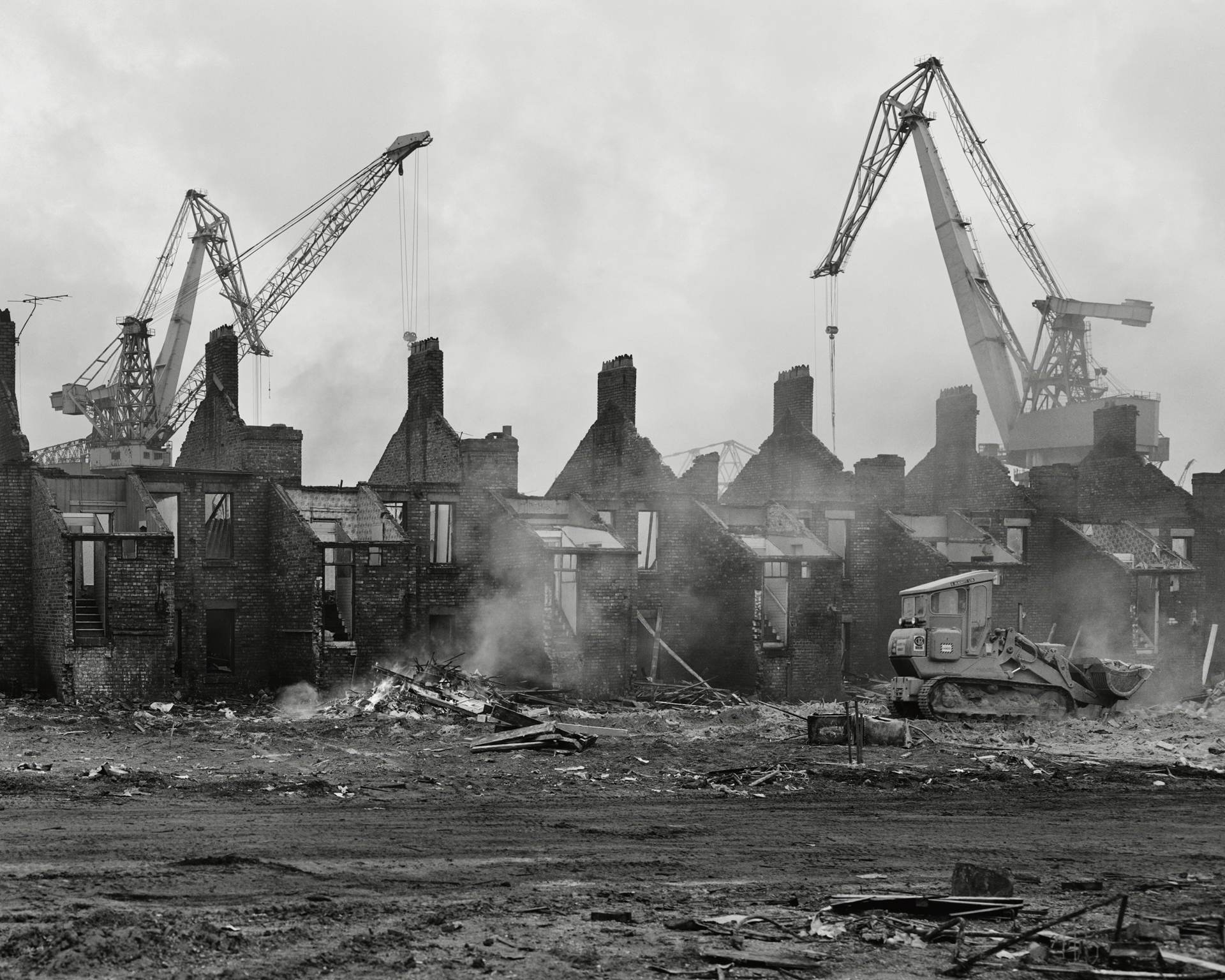 Black and white photograph of a row of terraced houses mid-demolition, their roofs stripped away and chimneys exposed, with two tall cranes and smoke rising behind them, taken in Wallsend, Tyneside, in 1976.