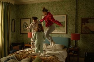 Image of a woman and two children jumping on an unmade bed. The headboard is blue and there are two orange lamps on either side of the bed. The room has green floral wallpaper all around it.