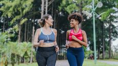 Two female friends running together in a forest – they are looking at each other and smiling wide. The lady on the left has her dark blonde hair up in a ponytail and is wearing a dark gray sports bra with white straps, gray leggings and a fitness watch on her left arm. The lady on the right has tightly cured brown hair and is wearing a pink and red chevron-patterned sports bra, navy blue leggings and a white fitness watch on her left arm.